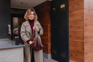 Modern yerleşim yerinin dışında bej renkli trençkot giyen bir kadın kahverengi deri çanta tutuyor. Sıcak gün ışığı desenli ahşap kaplama, gevşek duruş, yumuşak yüz ifadesi, mevsimsel görünüş, uygun günlük yaşam tarzı anı