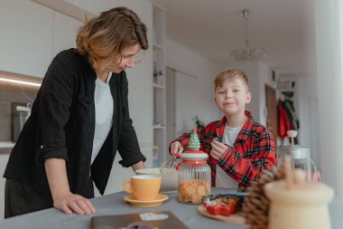 Anne, çerez kavanozunun yanında yemek hazırlarken küçük çocuğa yardım ediyor, sıcak ev sahnesi, yumuşak gün ışığı, rahat masa düzeneği, kırmızı ekose gömlek, canlı aile hayatı detayı ekliyor..