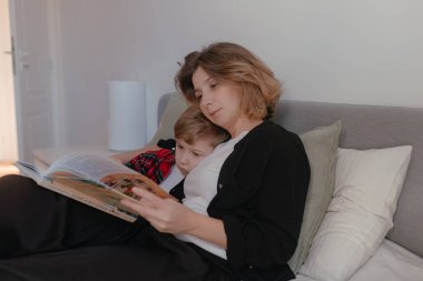 Doğal ışık yumuşak bir atmosfer yaratır. Annesi, çocuğun üzerine eğildiği bir kitabı tutar. Daha geniş açıdan çekilmiş sessiz okuma süresi, tarafsız iç mekandaki huzurlu aile etkileşimi..