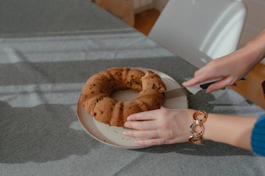 Sıcak gün ışığı mutfak sahnesi. Elleriyle halka şeklinde pasta kesiyor. Bıçak, kırıntı dokusu görünür yüzey, gri masa örtüsü yumuşak gölge deseni, basit ev yemeği hazırlama anı..