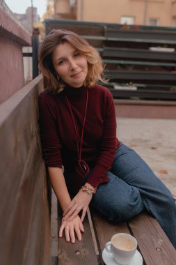 Yaşam tarzı portresi, tahtadan bir bankta oturan, öne doğru eğilip gülümseyen, bitişik kapuçino yakınlarında günlük giysiler giyen, ulaşılabilir şehir atmosferi yaratan bir kadın..