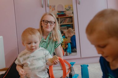 Kadınlar yarış pisti parçalarıyla oynayan çocukları gözlemler, ayna yüzeyi sahneleri, pastel mobilyalar odayı doldurur, oyuncaklar halının üzerine yerleştirilir aktif bir aile anı yaratır