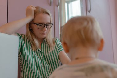 Gözlüklü kadın küçük çocuğun yanına eğilir, çizgili gömlek yumuşak gün ışığını yakalar, pastel dolap yüzeyi çiftin arkasına geçer, sakin duruş halı alanı boyunca aile aktiviteleri sırasında odaklanmış ruh hali yaratır.