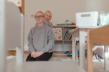 Yetişkinler oyun odası mobilyalarının yanında otururken, çocuklar sıcak aile odasında saç stili, yumuşak gün ışığı, mavi katmanlı elbise, çiçek saklama kutuları, tahta oyuncaklar, sahne arkasında kitap olan raflar..