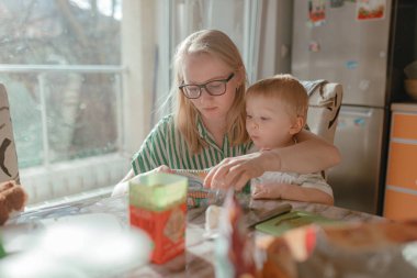 Yeni yürümeye başlayan bir kadın, aydınlık mutfağın içinde sıcak güneş ışığı, çizgili yeşil tişört, masanın yanında karışık yiyecek maddeleri ve konteynırlarla birlikte oturan bir çocuğun yemek paketini hazırlamaya yardım ediyor..