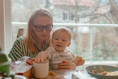 Anne, masa yüzeyine yakın yeni yürümeye başlayan çocuğu destekliyor, çocuk bisküviyle bardağa uzanıyor, büyük pencerenin karşısında yumuşak bir gün ışığı, çizgili tişört ayrıntıları, aile ortamında hassas bir hareketle yakalanan sıcak aile içi rutini..