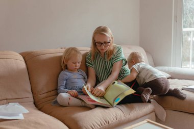 Yumuşak gün ışığı, anne masal kitabı okuyor. Yanında iki küçük çocuk, bej koltukta dinleniyor. Odaklanmış yüzler, rahat duruş, sıcak aile içi..