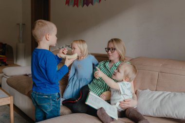 Gün ışığı, iki büyük çocuk yan yana durup muzlarını paylaşırken anneyi aydınlatıyor. Açık kitap yakınlarda dinleniyor. Sıcak iç mekanlarda enerjik bir aile etkileşimi..