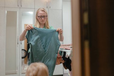 Çamaşır rutini sırasında büyük yeşil tişörtü değerlendiren genç bir kadın. Kurutma askılarının yanında düzgün sıralanmış giysiler, yumuşak iç aydınlatma, arkasında gardırop panelleri olan ev ortamları..