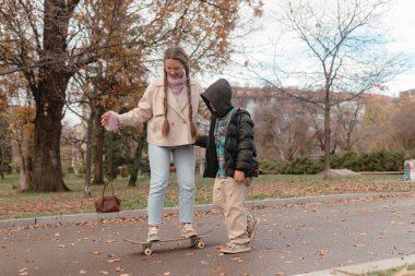 Kadın kaykaya binerken, oğlan sonbahar antrenmanı sırasında açık hava parkında asistanlık yapıyor. Arkaplan ağaçları ve binalar etkin öğrenme anını çerçevelerken kuru yapraklar kaldırımı kapatır.