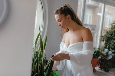 Kapalı duran uzun yılan bitkisi yumuşak bir şekilde yuvarlak pencerenin yakınındaki yapraklara dokunuyor. Gün ışığı sakin bir iç mekan yaratıyor. Beyaz giysi az temiz estetik ekliyor..