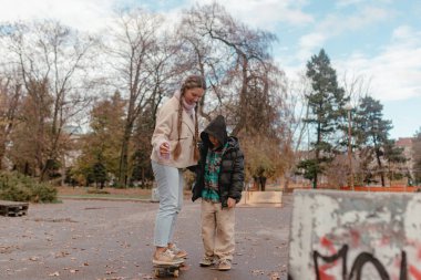 Yan gören kadın gülümsüyor, el uzatılmış kılavuz hareketi, çocuk odaklı kaykay duruşu, bej ceket pembe eldivenler, siyah kabarık ceket, park ağaçları arka planı, destekleyici ebeveynlik sahnesi