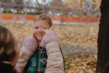 Kadın ellerini çerçeveye koyarken çocuk gülümsüyor. Sonbahar parkı arka planı, sarı yapraklar, sığ odak, samimi aile portresi, nazik şakacı etkileşim