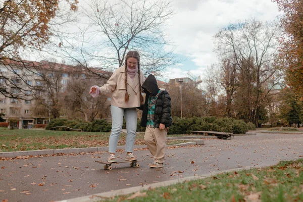 Genç kadın bej ceketli açık renk kot, ayakta duran kaykayı destekliyor. Sonbahar parkı patikası, düşen yapraklar, şehir binaları arka plan, bulutlu gökyüzü, açık havada aile aktiviteleri.