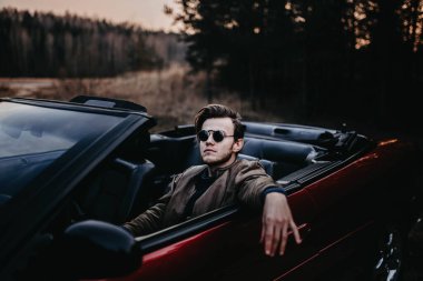  Sakallı vahşi bir adam doğada üstü açık kırmızı güneş gözlüklü bir arabada oturuyor. Erkek bekar ve sürücü tarzı.