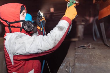 Solunum cihazındaki erkek işçi, arabanın alt kısmına korozyon önleyici bir boyayla kompresör uyguluyor..