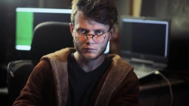 tired hacker in a black room looking at the camera and computers with several screens in the background 