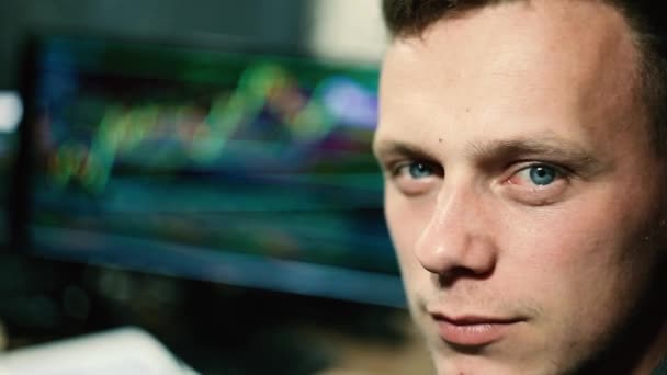 一个年轻的斯拉夫男性黑客用股票交易所和图表对着电脑 看着相机和眨眼的镜头 — 图库视频影像