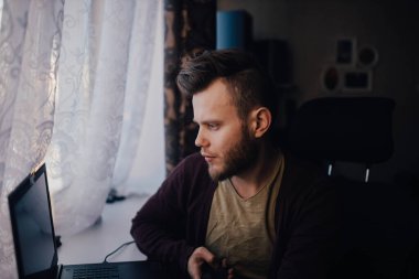 Sakallı adam evinde oturuyor ve dizüstü bilgisayara bakıyor.