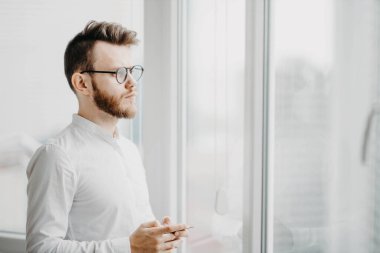 Erkek işadamı pencerenin kenarında durup beyaz gömlekli, sakallı genç bir adam gibi düşünüyor.