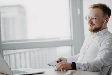 Gözlüklü, kablosuz kulaklık takan genç adam dizüstü bilgisayarla ve video konferansında çalışıyor.