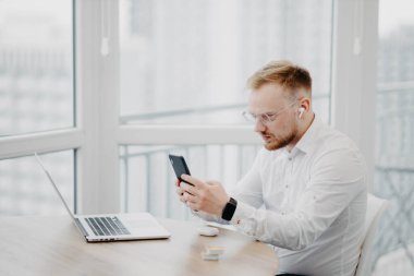 Gözlüklü, kablosuz kulaklık takan genç adam dizüstü bilgisayarla ve video konferansında çalışıyor.
