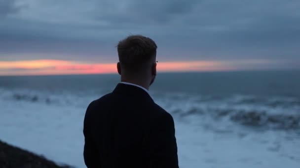 日落时 身穿外套的寂寞男子站在海滨 激荡着海浪 — 图库视频影像
