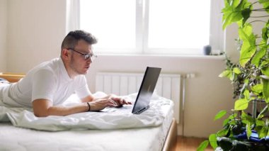 Genç adam beyaz bir yatakta yatıyor ve evinde saksıların ve bitkilerin arasında dizüstü bilgisayarla çalışıyor.