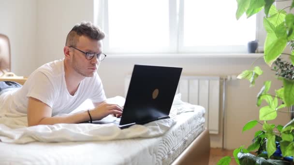 Młody Człowiek Leży Białym Łóżku Pracuje Laptopem Okularach Wśród Doniczek — Wideo stockowe
