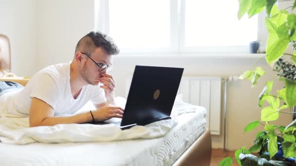 Młody Człowiek Leży Białym Łóżku Pracuje Laptopem Okularach Wśród Doniczek — Wideo stockowe
