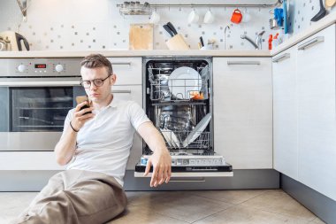 Adam akıllı telefondan bulaşık makinesinin talimatlarını okuyor, kocası da mutfakta ev işi yapıyor.