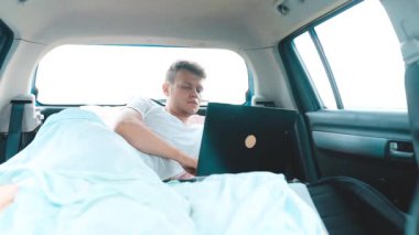 Dizüstü bilgisayarda çalışan bir adam arabada battaniyenin içinde yatıyor. 