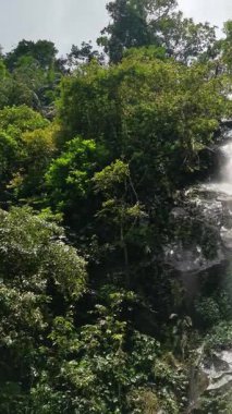 This footage shows a beautiful waterfall cascading down a rocky slope surrounded by dense, green foliage. Experience the serene beauty of nature with this captivating scene, ideal for travel.