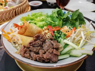 Izgara sığır eti, çıtır börek, taze otlar, marul, fasulye filizi ve düzgün bir şekilde hazırlanmış sebzeler içeren renkli bir Vietnam tabağı..