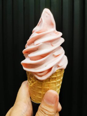 Bir el, üzerinde pembe servis dondurması olan waffle külahını tutar, parlak kıvrımları koyu çizgili bir arka plana karşı durur, tatlılığı ve neşe dolu hoşgörüyü çağrıştırır.