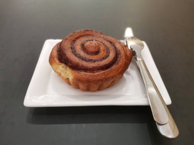 Altın kabuklu ve zengin spiral dolgulu yeni pişmiş bir tarçın rulosu bir tabakta duruyor, çatal bıçaklarla eşleştiriliyor, hoş ve davetkar bir kahvaltı veya tatlı anı yaratıyor.