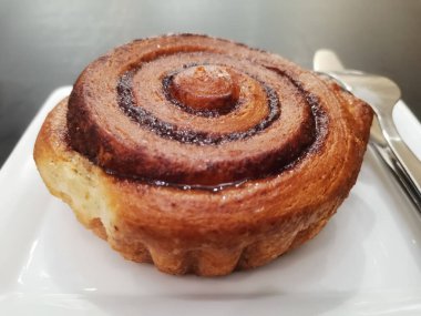 Sıcak tarçınlı rulo, parlak jöleli ve zengin spiral dolgu bir tabakta duruyor çatal ve bıçakla eşleştirilmiş sıcak bir kahvaltıyı veya tatlıyı tatlılıkla dolu bir tatlı anına davet ediyor.