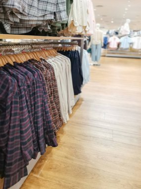 Ahşap zeminli parlak bir perakende giyim mağazasında asılı duran çeşitli ekose ve katı renkli gömlekler, modern bir alışveriş ortamında alışveriş yapanlara çeşitli moda seçenekleri sunuyor..