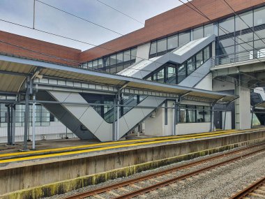 Tangerang, Banten, Endonezya, 1 Aralık 2025. Jatake İstasyonu inşaatta. İçinde cam duvarlar ve yürüyen merdivenler olan modern bir istasyon binasının yanında boş bir demiryolu platformu. Tren rayları bulutlu bir gökyüzü altında platforma paralel gider..