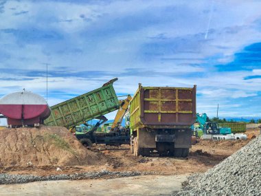 Bogor, Endonezya, 5 Aralık 2025. İki büyük damperli kamyon ve bir kazıcı mavi bulutlu bir gökyüzünün altında bir inşaat alanında çalışıyor. Bir kamyon eğilmiş, toprak ve çakıl yükünü boşaltıyor..