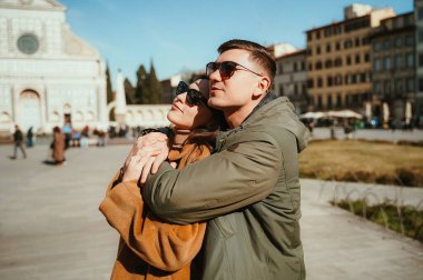 Güneşli bir meydanda şefkatle kucaklaşan bir çift tarihi mimari ile çevrili, sevgi ve şefkat dolu bir anı yakalıyorlar. Her iki birey de günlük kış kıyafetleri ve güneş gözlüğü takıyor.