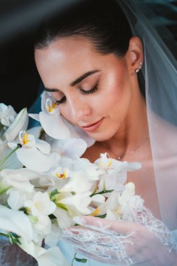 Gelin nazikçe bir buket beyaz orkide tutuyor, gözleri kapalı, sakin bir ifadeyle anı kucaklıyor. Dantel eldivenleri ve peçesi sahneye biraz zarafet katıyor..