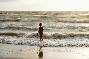 Çocuk plajdaki yumuşak dalgalarda oynar, gün batımında okyanusun sakinleştirici varlığını kucaklar. Doğayla olan bağımızı ve çocukluk sevincimizi yansıtan huzurlu bir an..
