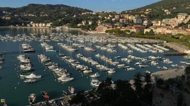 Renkli bir sahil kasabasının arka planında yer alan sayısız tekneyle dolu hareketli bir limanın manzarası. Kasabayı çevreleyen tepeler manzaralı ve huzurlu bir atmosfer sunuyor..