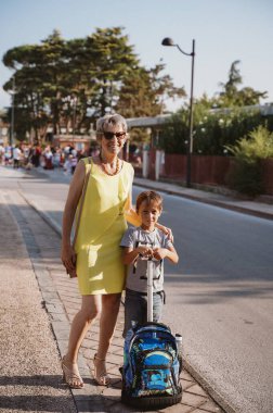 Güneşli bir sokakta elinde renkli bir sırt çantası tutan genç bir çocukla gülümseyen yaşlı bir kadın. İfadeler neşe ve bağlantı gösteriyor, aile sıcaklığını ve desteğini vurguluyor