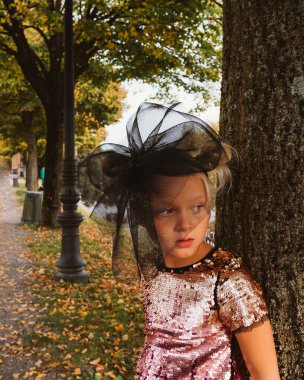 Işıl ışıl pullu elbisesi ve büyük siyah şapkası olan küçük bir çocuk bir ağaca yaslanmış, yana bakıyordu. Sonbahar yaprakları ve sokak lambaları atmosferik bir zemin oluşturur.