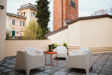 Modern beyaz sandalyelerin ve turuncu bir masanın yer aldığı açık teras tarihi mimari ve yeşilliğin arka planına karşı kurulmuş, rahatlatıcı ve davetkar bir atmosfer yaratıyor.