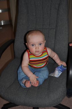 Meraklı bir yüz ifadesi olan bebek gri bir sandalyede elinde renkli bir kağıt parçasıyla oturuyor. Çizgili bir bluz ve kot pantolon giyen bebek dikkatle kameraya odaklanıyor..