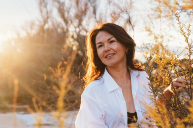 Beyaz gömlekli kadın doğanın ortasında gülümsüyor altın saat güneş ışığı içinden süzülüyor, sıcak ve sakin bir ortam yaratıyor. Yüzü rahat ve neşeli..