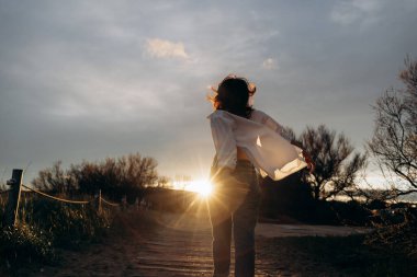 Kırsal bir yolda gün batımına doğru enerjik bir şekilde koşan kişi, rüzgarla savrulan bir gömlekle rüzgarda savruluyor. Güneş ışığı parlıyor, yolu aydınlatıyor ve sıcak, huzurlu bir atmosfer yaratıyor..